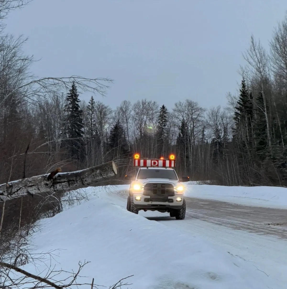 Rig Dawgs pilot truck head-on at dusk, amber beacons and D sign lit, headlights on, snow road